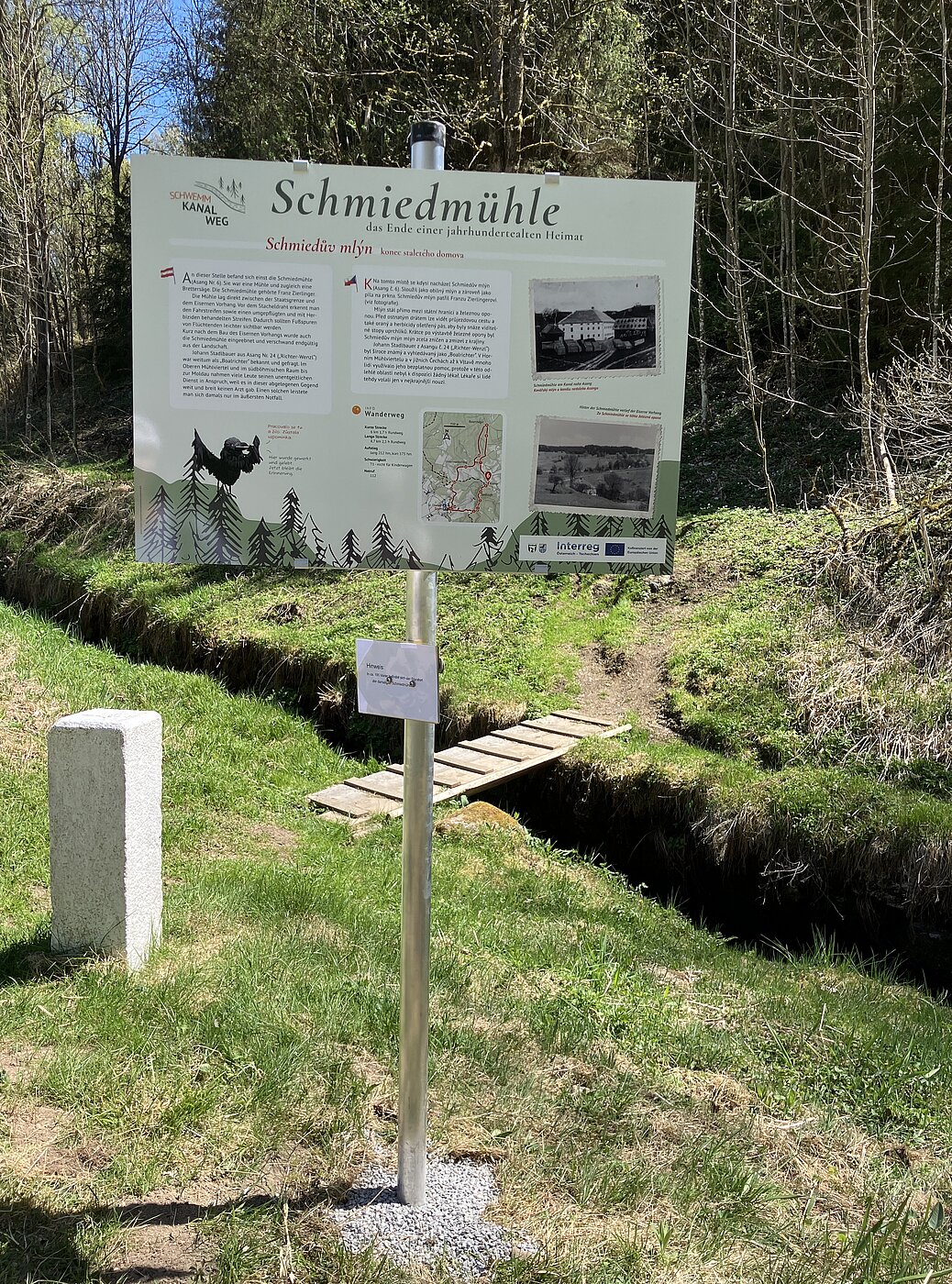 Person steht am Waldrand an einem Bach und steuert mit langem Stock Holzstücke im Wasser; im Hintergrund dichter Nadelwald